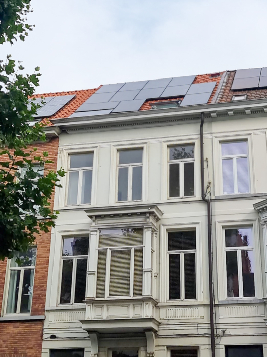 Stadswoning met witte gevel en rode dakpannen, waarop zwarte zonnepanelen geplaatst zijn boven meerdere dakkapellen