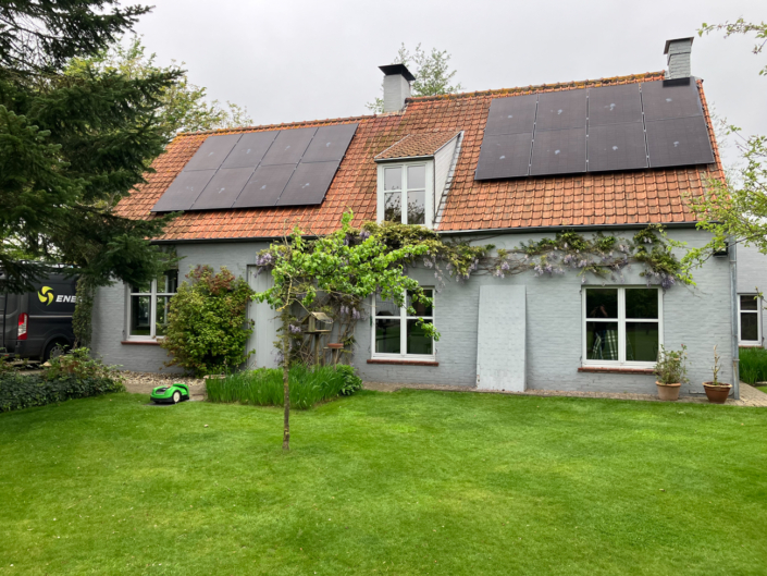 Zonnepanelen op oranje dakpannen van landelijke woning met blauwe ramen en veel groen